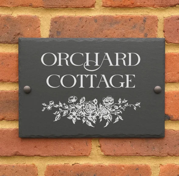 Personalised engraved slate house sign reading Orchard Cottage with a floral design, mounted on a brick wall